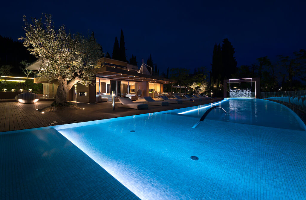 A nighttime photo of the exterior of a brightly lit luxury home with a large pool in the backyard.