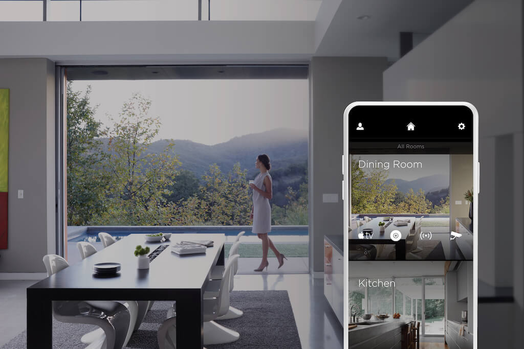 A photo of a woman walking through her dining room in front of a massive window wall displaying the mountains behind her.