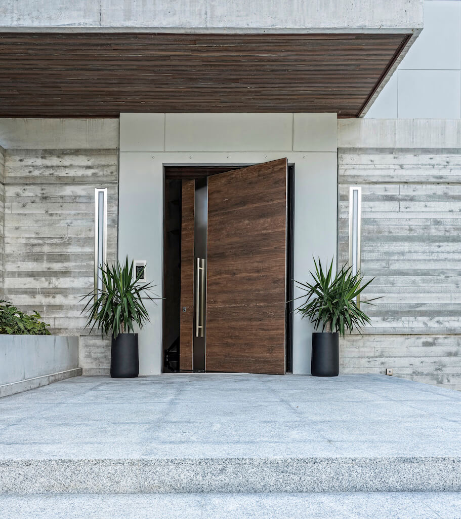 A photo of an oversized pivot door covered in wood and stainless steel at the front entrance of a luxury home.